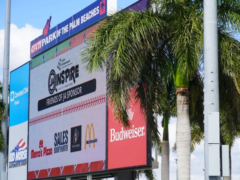 JA Inspire sponsorship displayed on the CACTI Park of the Palm Beaches scoreboard, showing Friends of JA Sponsor logos including Cleveland Clinic Florida, Budweiser, Marco's Pizza, and McDonald's