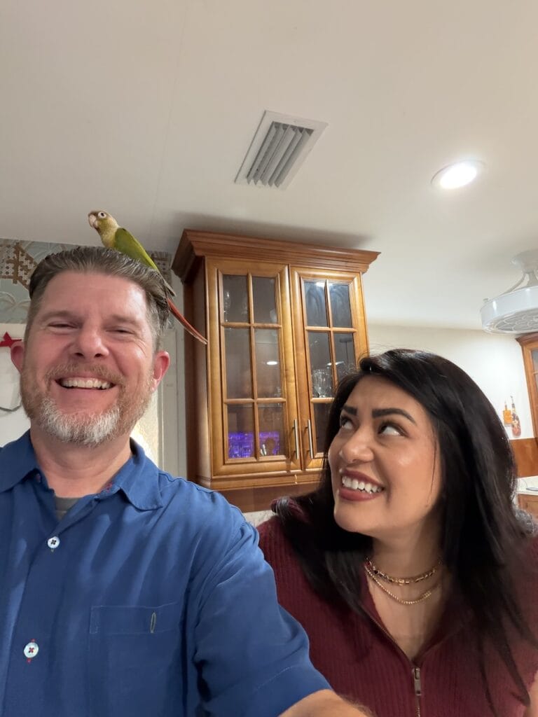 John W. Brandes and a woman smiling indoors, with a small green parrot perched on his head