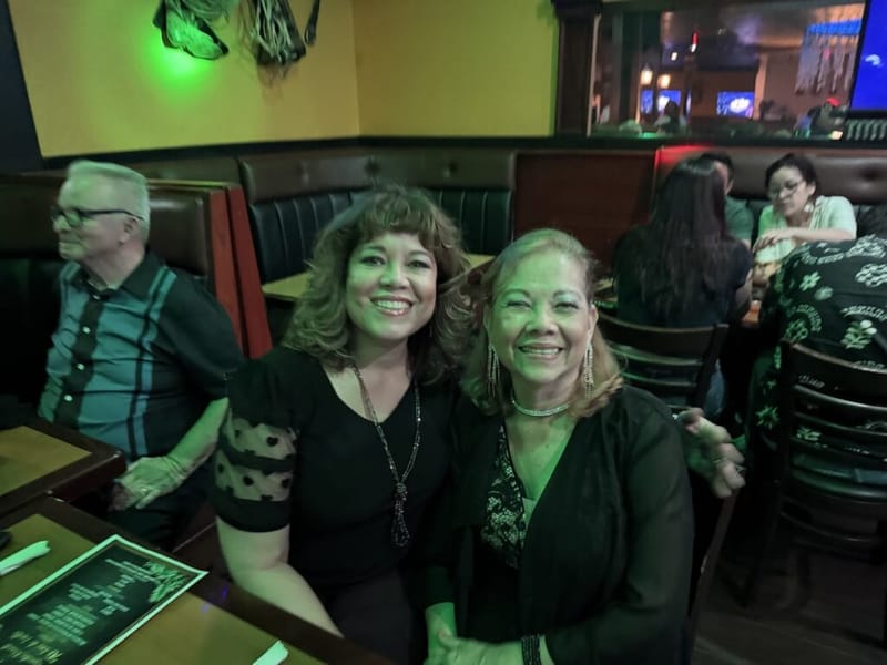 Two women smiling together at a booth inside Rancho Nando restaurant, both wearing black, with the dining room visible behind them