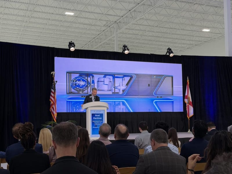 Noel Martinez, President of Palm Beach North Chamber of Commerce, speaks at the Ampera grand opening event, with a large product rendering displayed on screen behind him and an audience seated in the foreground