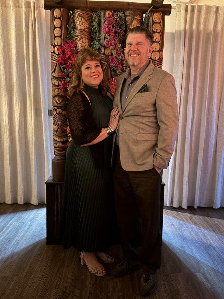 A couple dressed for a holiday party, posing in front of a carved tiki-style wooden backdrop decorated with floral garlands