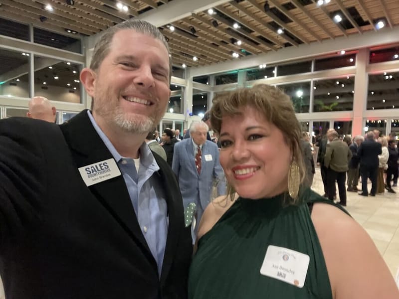 John Brandes and Ana Brandes smiling together at a holiday party, both wearing name badges, in a large event venue with guests in the background