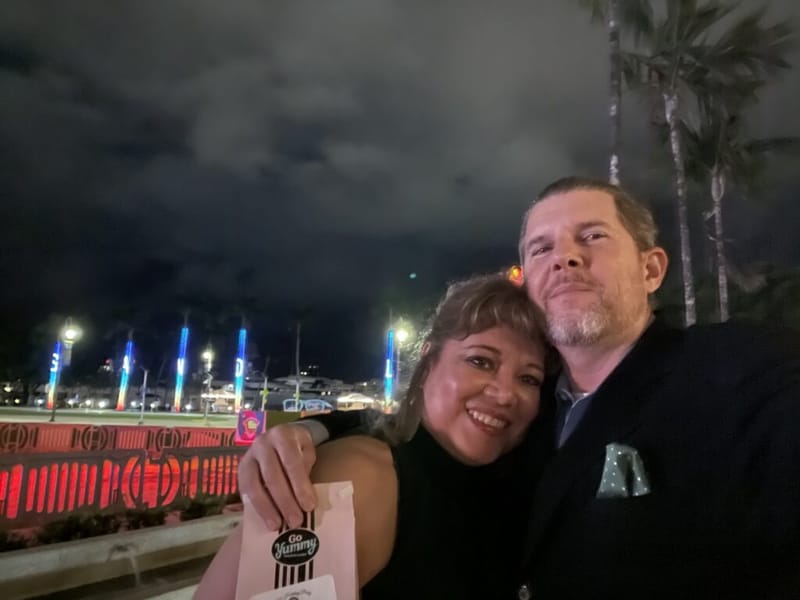 John W. Brandes and a companion at an outdoor holiday party at night, colorful event lights and palm trees behind them, holding a Go Yummy gift bag